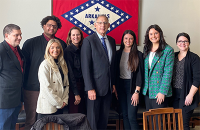 Eight people standing in front of the Arkansas state flag in the senators office.
